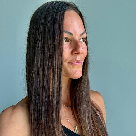 Danielle with brown eyes and brown straightened chest-length hair standing against a gray background, wearing a black v-neck tank top with crystal necklace